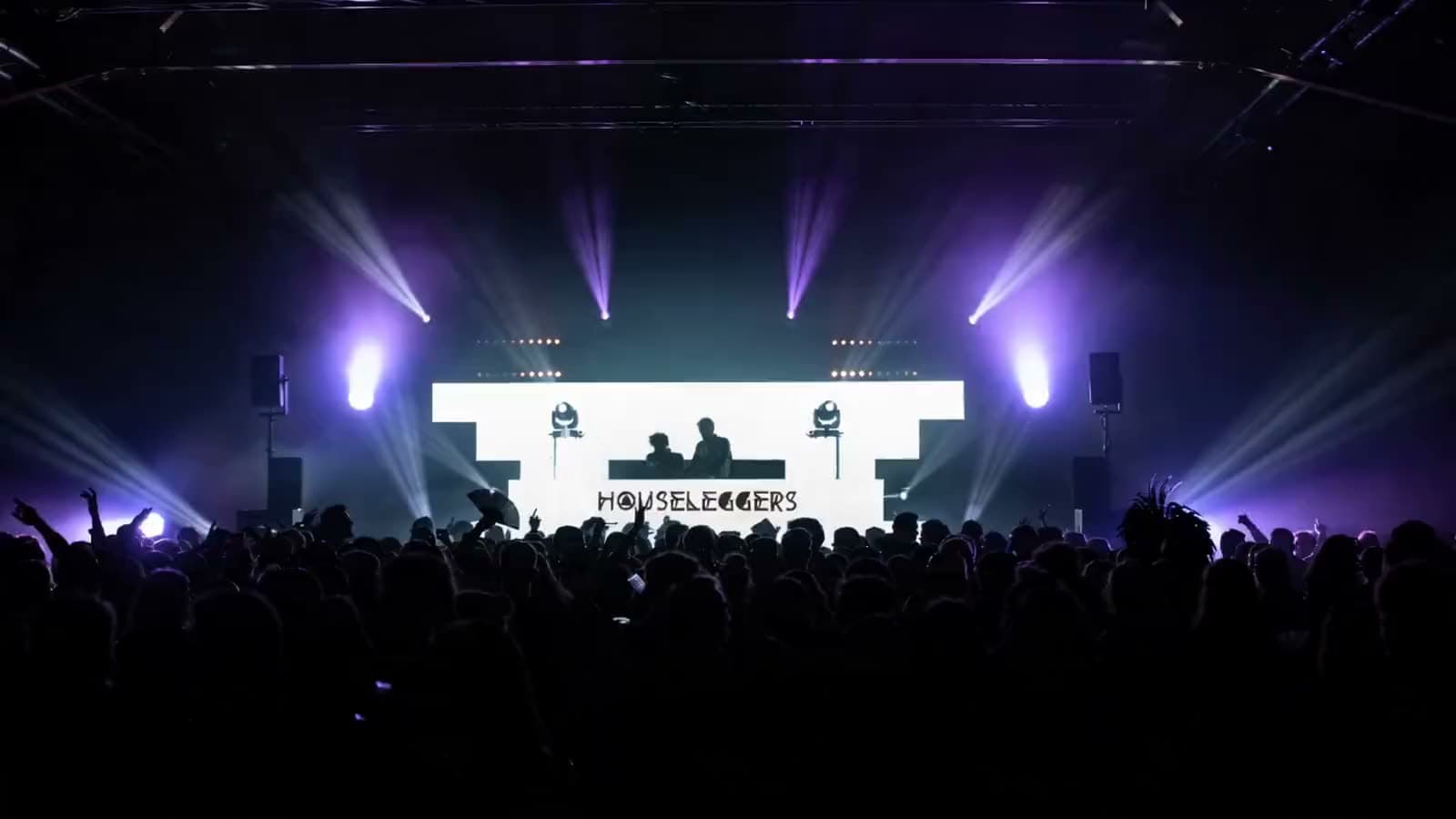The Houseleggers on the decks in front of a festival crowd, violet beams sweeping above the audience